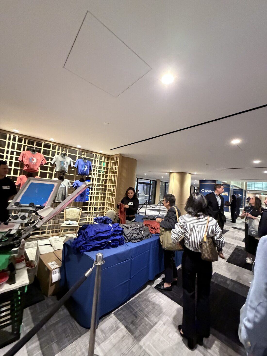 Guests waiting at an indoor lobby apparel station while a crew member sorts folded shirts, with printed samples displayed on the wall behind