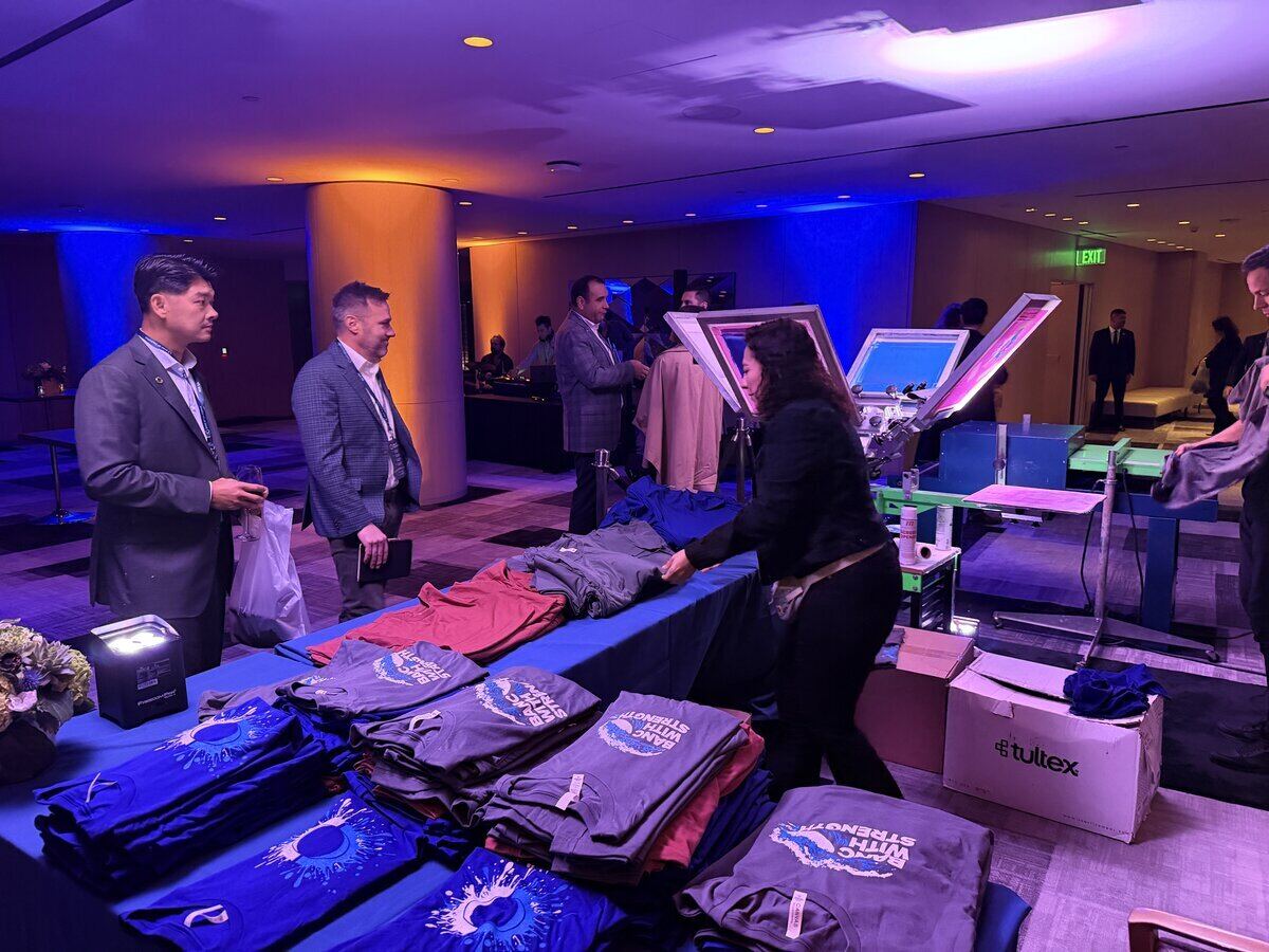 Live apparel station mid-service at an indoor evening event, folded printed shirts staged in colour-sorted stacks beside the press
