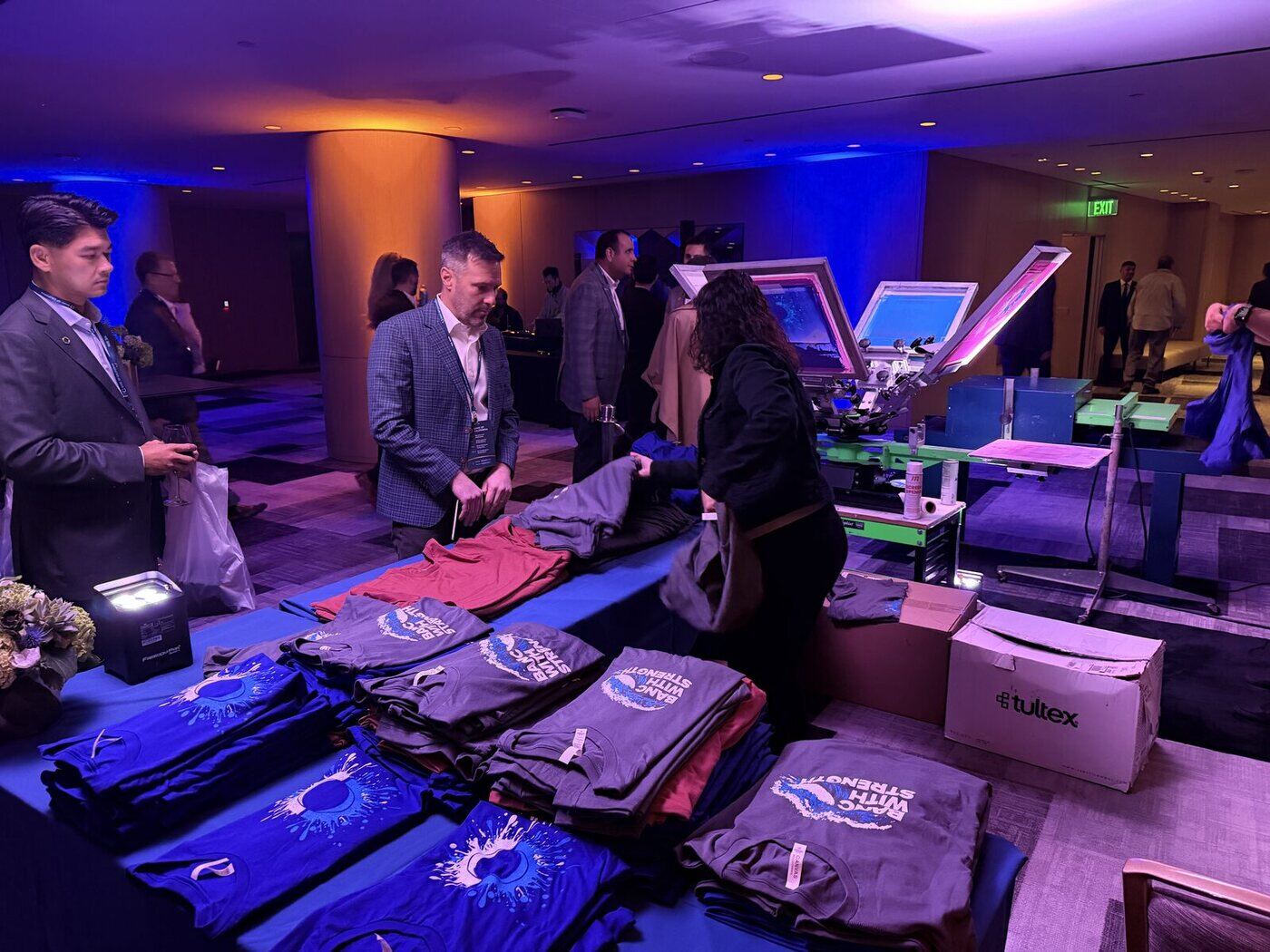 Evening corporate reception with a live apparel station: stacks of folded printed shirts on a draped table while a crew member works a manual press behind them