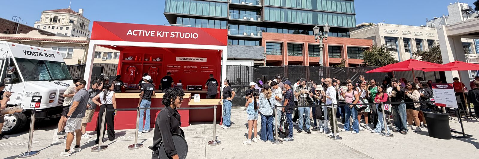 Red outdoor kit studio booth on a downtown plaza, with a personalization counter, locker bays of jerseys behind it and a long stanchioned guest queue running across the concrete