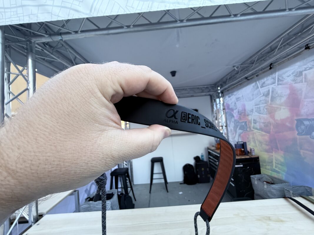 Freshly engraved strap held up in a tented event studio during setup, with the work table and staging bins behind it